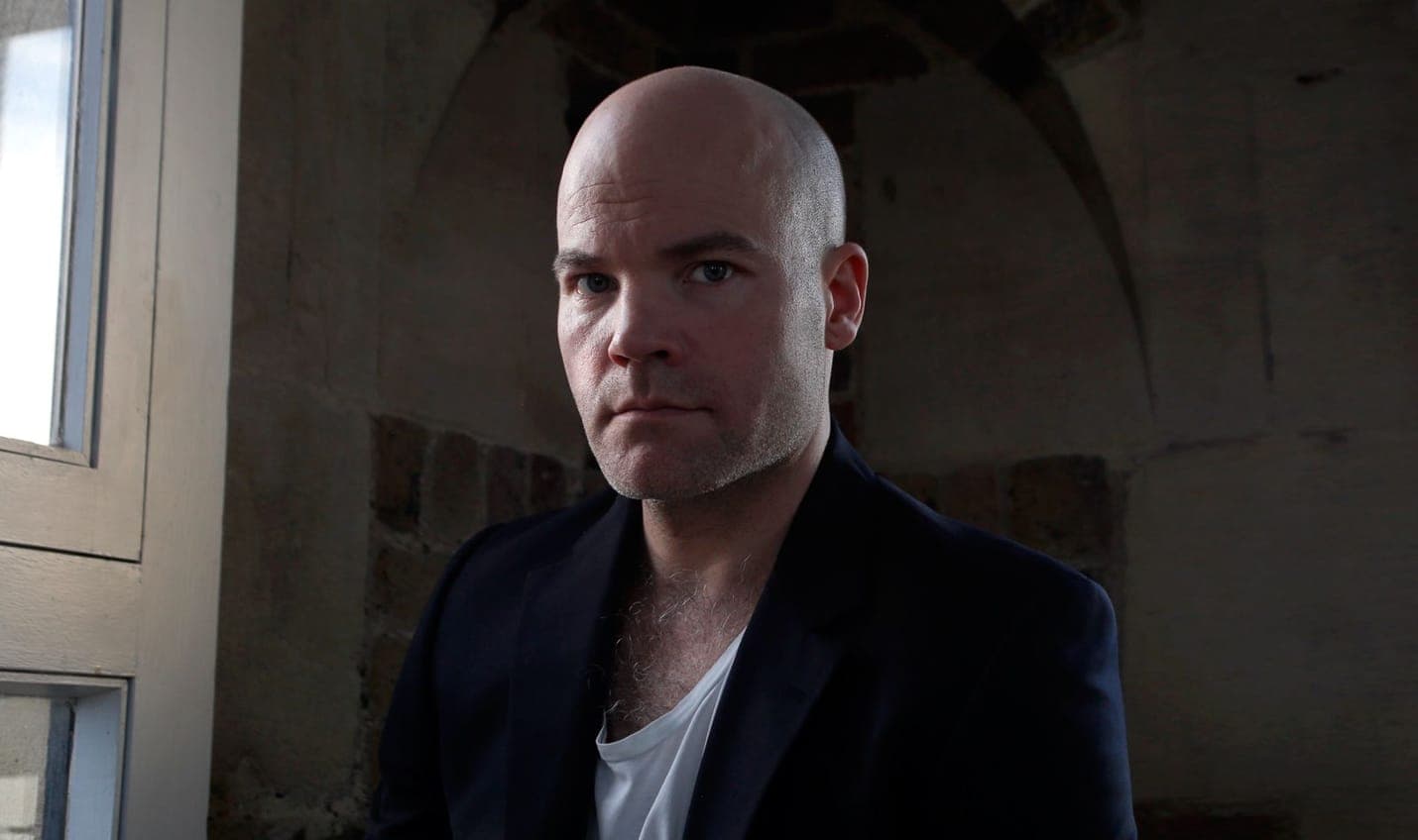 Henrik Delehag con chaqueta oscura y camiseta blanca, iluminado por una ventana a su izquierda, ante el arco de piedra desnuda de su estudio en la torre